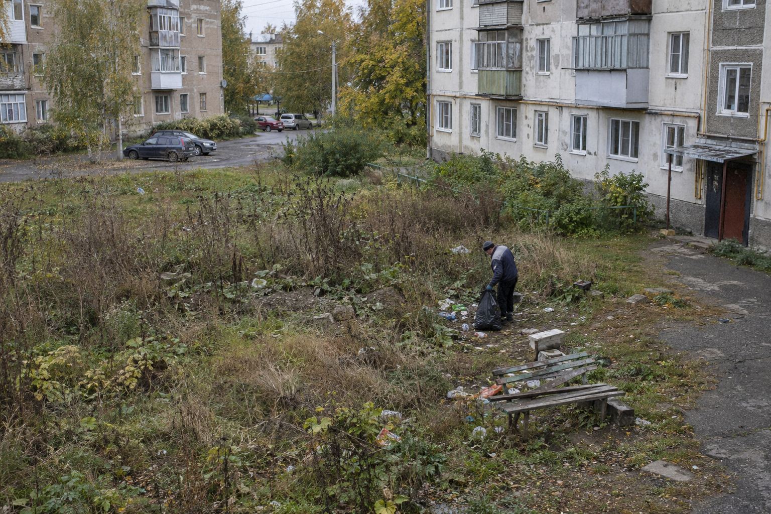 Заросший пустырь во дворе пятиэтажек, один человек собирает мусор в мешок